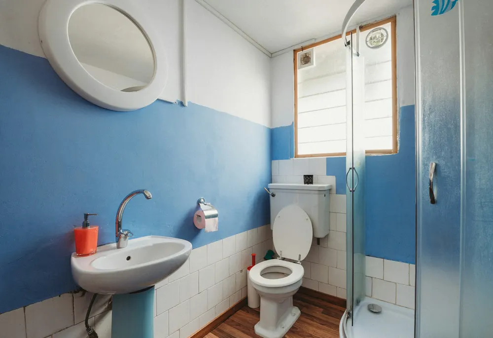 The test bathroom before: half-blue painted walls over white tiles, a round mirror, a pedestal sink, a toilet and a glass shower