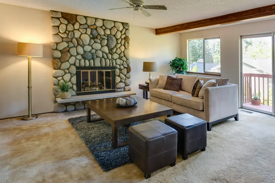 Before: 1970s living room with a river-rock fireplace, beige carpet, a light sofa and two leather ottomans