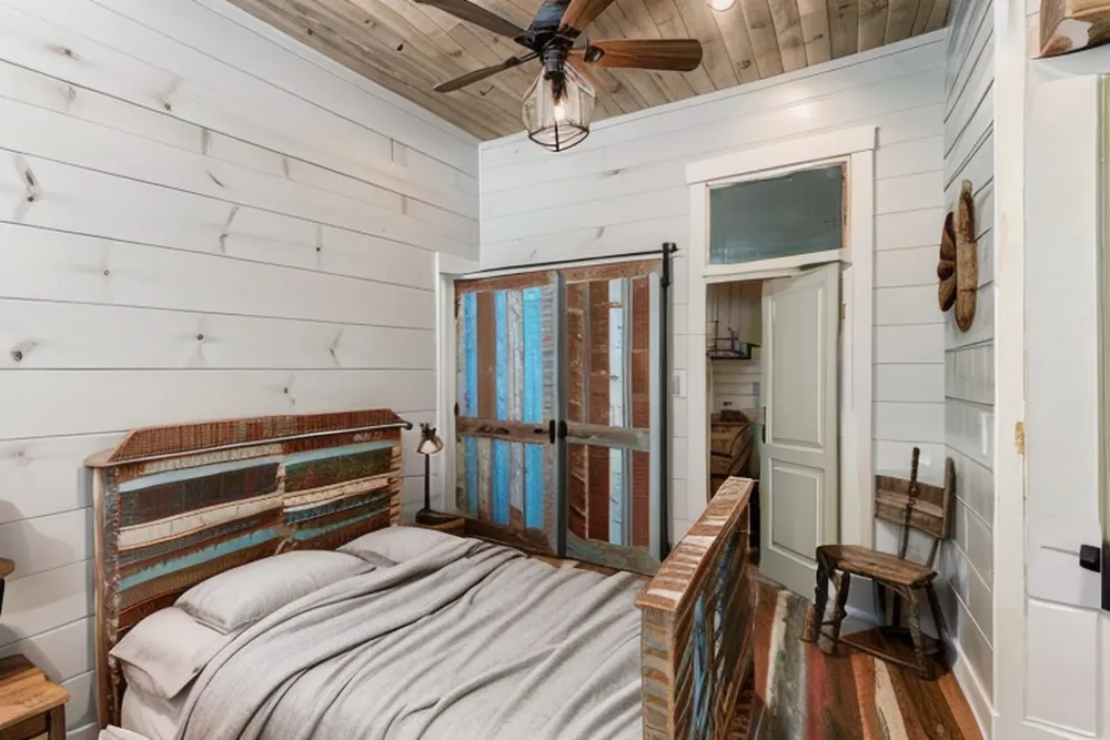 After: the same bedroom with white shiplap walls, a painted reclaimed-plank bed, barn-style wardrobe doors and a fan with a lantern light