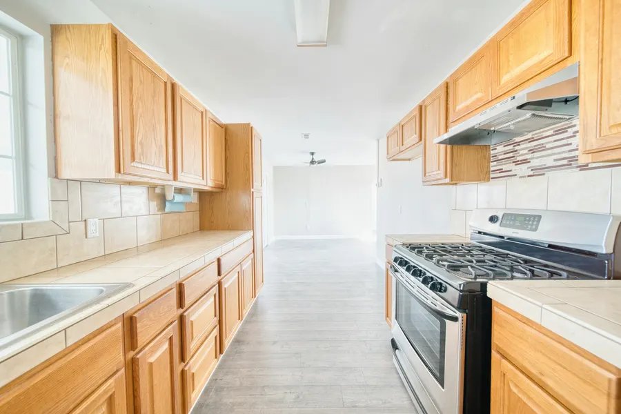 Before: empty galley kitchen with honey-oak cabinets, beige tile counters and a steel gas range