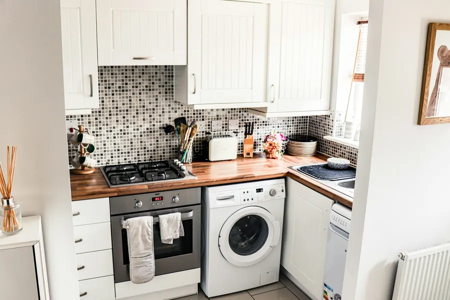 Before: small kitchen with white cabinets, a wood worktop, grey mosaic backsplash and a washing machine