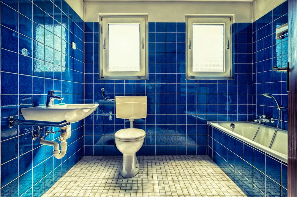 Before: a 1970s bathroom tiled in bright blue with two windows, a wall-hung sink, a toilet and a tub