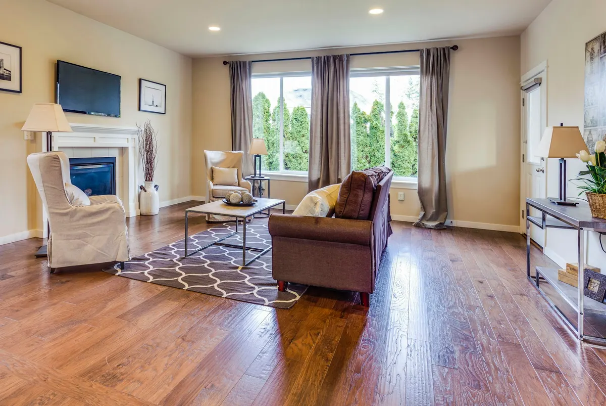 The test room before: a beige living room with a white fireplace under a wall TV, two windows with grey curtains, a brown loveseat and a dark wood floor